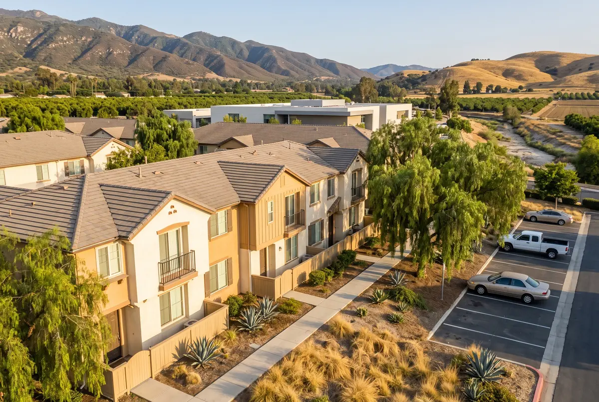 Illustration of two-story attached townhomes with small balconies along a shared landscaped walkway, with a low college building and rolling hills beyond.
