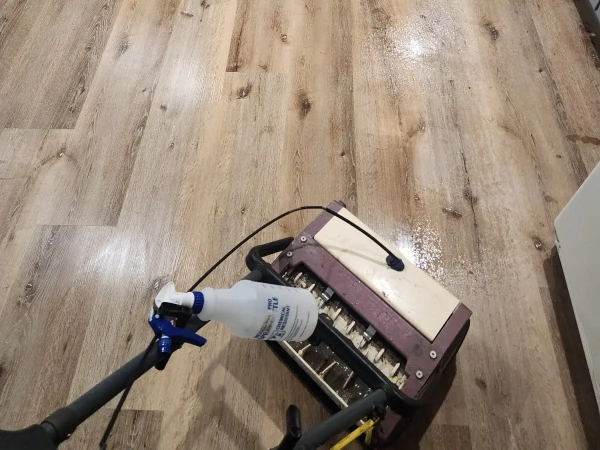 Photo of a wood-look plank kitchen floor being scrubbed by a cylindrical brush machine, with a spray bottle of cleaning solution on the handle and gray cabinets beyond.